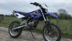 Moto sherco 50cc supermotard bleue et violette vue avant gauche 3 4 sur béquille chaine moteur échappement fourche jantes à rayons pneus route gravier prairie cloture arbres ciel nuageux éclairage naturel
