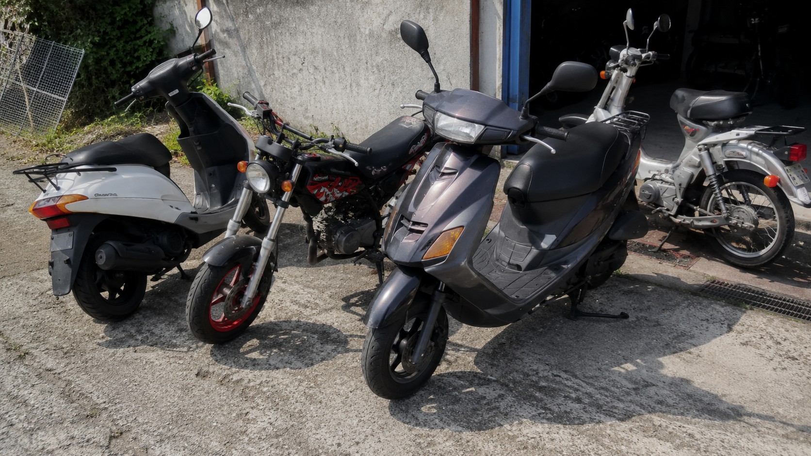 Suzuki 50cc d'occasion exposés en extérieur au soleil, scooter gris au premier plan, scooter blanc, moto type Street Magic noire et rouge avec jantes rouges, cyclomoteur gris en arrière-plan, carrosseries et éléments mécaniques visibles, comparaison de modèles 50cc et prix du marché en 2026