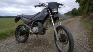yamaha_dt_50cc_noire_enduro_supermotard_stationnee_sur_chemin_de_gravier_vue_trois_quarts_avant_gauche_cadre_tubulaire_moteur_2_temps_pot_detente_fourche_telescopique_jantes_a_rayons_pneu_route_frein_a_disque_avant_garde_boue_haut_phare_rectangulaire_selle_noire_campagne_francaise_ciel_couvert_lumiere_naturelle_photo_documentaire_realiste_2026