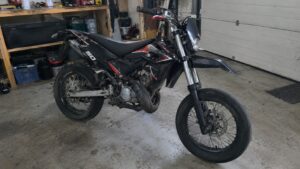 Moto 50cc supermotard noire, rouge et blanche, vue trois-quarts avant, roues à rayons et pneus route, dans un garage