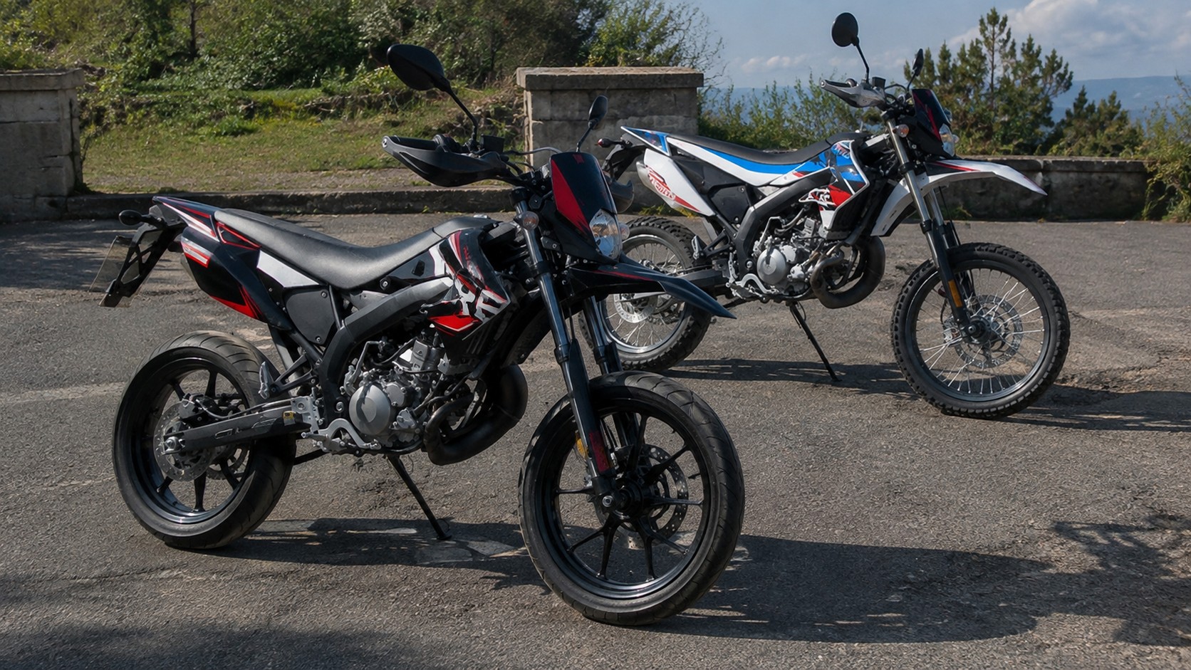 Deux Derbi Senda Xtreme 50cc stationnées sur une route asphaltée en plein soleil, version SM supermotard au premier plan avec jantes alliage noires, pneus route, frein à disque avant, fourche télescopique, carénages noir rouge blanc et moteur 50 cm³ apparent, version R enduro à l’arrière avec roues à rayons, pneus tout-terrain à crampons, garde-boue avant surélevé, béquille latérale, carénages blanc bleu rouge, muret en pierre et végétation verte en arrière-plan, vue trois-quarts avant, photographie, lumière naturelle directe