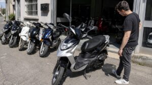 Devanture_de_magasin_50cc_en_plein_soleil_plusieurs_scooters_alignes_a_exterieur_et_dans_le_showroom_client_en_tenue_noire_inspectant_un_scooter_noir_et_gris_stationne_sur_bequille_centrale_vue_grand_angle_documentaire_16_9