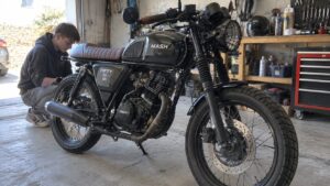 Mash Fifty 50 café racer 50cc noire, selle marron nervurée, réservoir MASH, moteur monocylindre, échappement bandé, roues à rayons, frein à disque avant, phare grillagé, soufflets de fourche, adolescent accroupi près de la roue arrière dans un garage mécanique éclairé par lumière naturelle