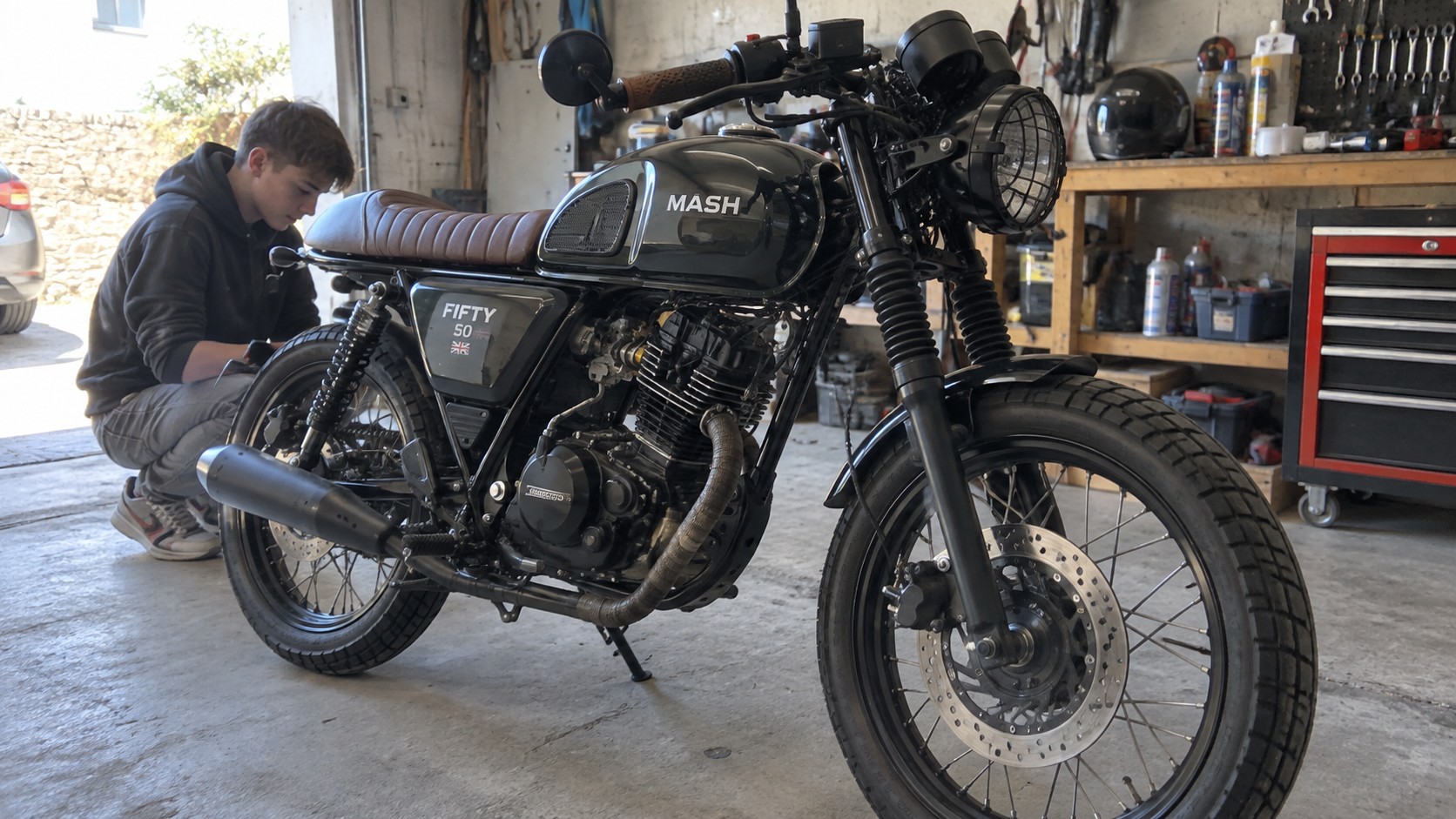 Mash Fifty 50 café racer 50cc noire, selle marron nervurée, réservoir MASH, moteur monocylindre, échappement bandé, roues à rayons, frein à disque avant, phare grillagé, soufflets de fourche, adolescent accroupi près de la roue arrière dans un garage mécanique éclairé par lumière naturelle