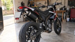 supermotard 50cc noir vue arrière trois quarts dans garage, double silencieux échappement superposés côté gauche, embouts métalliques, roue arrière avec disque de frein, bras oscillant, amortisseur central, plaque arrière et feu rouge, carénages noirs rouges blancs, atelier mécanique en arrière-plan, lumière naturelle, 2026