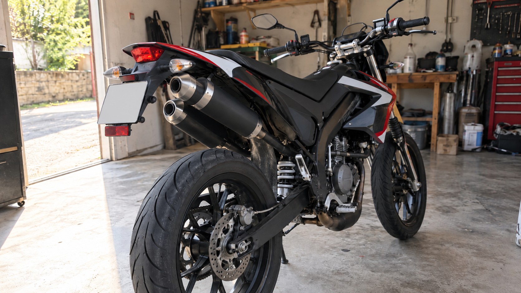 supermotard 50cc noir vue arrière trois quarts dans garage, double silencieux échappement superposés côté gauche, embouts métalliques, roue arrière avec disque de frein, bras oscillant, amortisseur central, plaque arrière et feu rouge, carénages noirs rouges blancs, atelier mécanique en arrière-plan, lumière naturelle, 2026