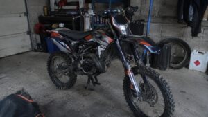 Moto enduro 50cc noire, orange et blanche, vue trois-quarts avant, sur béquille latérale dans un garage, pot de détente et pneus à crampons visibles
