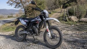 Moto enduro 50cc homologuée blanche noire et rouge, modèle exact non identifiable visuellement, moteur 2 temps avec pot de détente, fourche télescopique, frein à disque avant, roues à rayons et pneus tout-terrain à crampons, béquille latérale, rétroviseurs et support de plaque, stationnée sur chemin rural caillouteux en bord de route, arbres et relief montagneux en arrière-plan, guide enduro 50cc 2026
