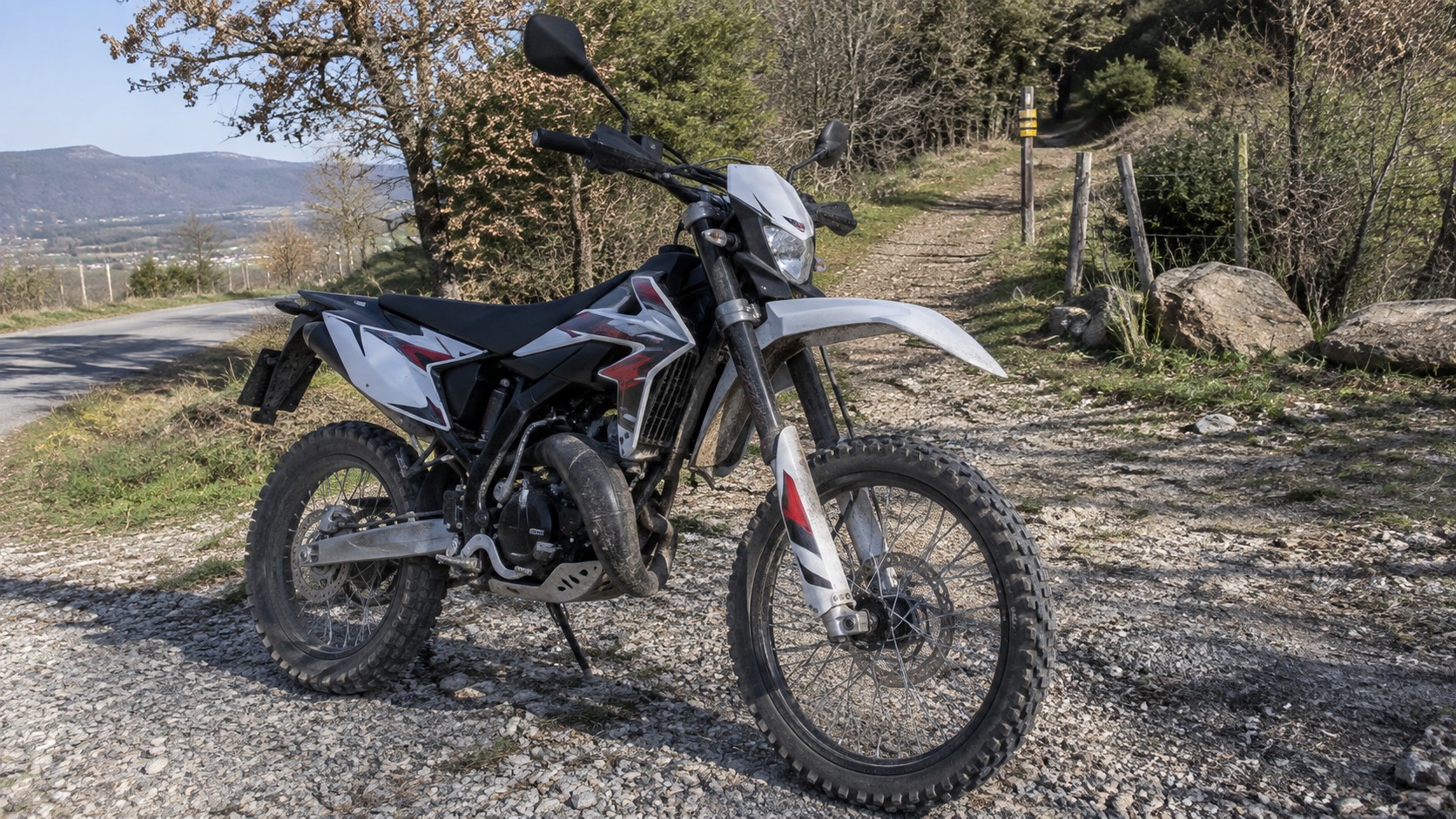 Moto enduro 50cc homologuée blanche noire et rouge, modèle exact non identifiable visuellement, moteur 2 temps avec pot de détente, fourche télescopique, frein à disque avant, roues à rayons et pneus tout-terrain à crampons, béquille latérale, rétroviseurs et support de plaque, stationnée sur chemin rural caillouteux en bord de route, arbres et relief montagneux en arrière-plan, guide enduro 50cc 2026