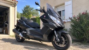Maxi scooter 50cc noir gris vue trois quarts avant gros carénage bulle teintée double selle guidon rétroviseur roues disques frein avant échappement garage maison française allée gravier lumière naturelle soleil ombres naturelles