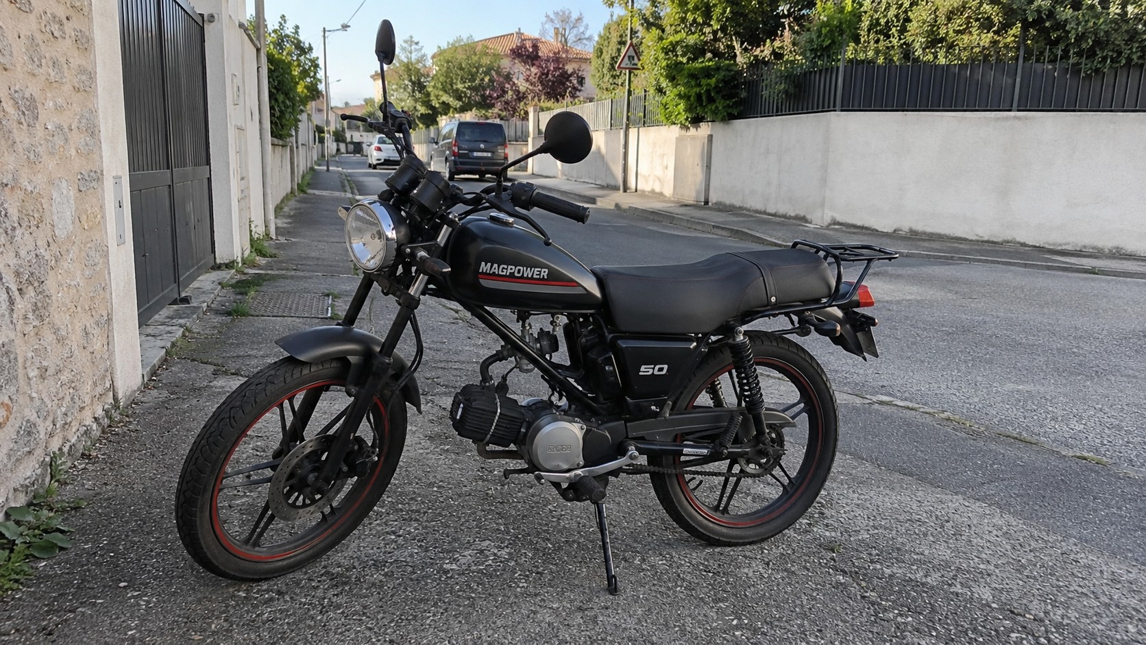 Magpower 50cc noire stationnée sur béquille latérale dans rue résidentielle ensoleillée, vue trois-quarts côté gauche, réservoir noir avec marquage MAGPOWER et filet rouge, selle longue noire, porte-bagages arrière, phare avant rond cerclé de chrome, deux rétroviseurs noirs, moteur 50 cc apparent, échappement et transmission visibles, fourche avant noire, frein à disque avant, jantes à bâtons noires avec liseré rouge, pneu arrière large, murs blancs et portail sombre à gauche, trottoir et chaussée grise, voitures garées en arrière-plan, arbres et végétation au-dessus des clôtures, panneau triangulaire de signalisation au fond, lumière naturelle directe et ombres nettes, photographie documentaire grand-angle 2026