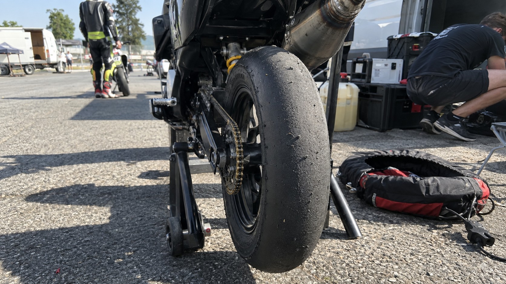 pneu slick arrière de moto 50cc de piste, gomme sans sculptures marquée par roulage et dépôts de caoutchouc, roue arrière levée sur béquille de stand, transmission par chaîne et couronne visibles, échappement sous selle, paddock de circuit en plein soleil