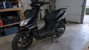 scooter_rmax_50cc_noir_vue_trois_quarts_avant_garage_francais_porte_sectionnelle_établi_outils_bacs_rangement_sol_béton_fourche_avant_frein_à_disque_roue_arrière_transmission_moteur_thermique_selle_noire_repose-pieds_carénage_sportif_autocollants_rmax_50cc_lumière_naturelle_ambiance_atelier_mécanique_2026_09_29