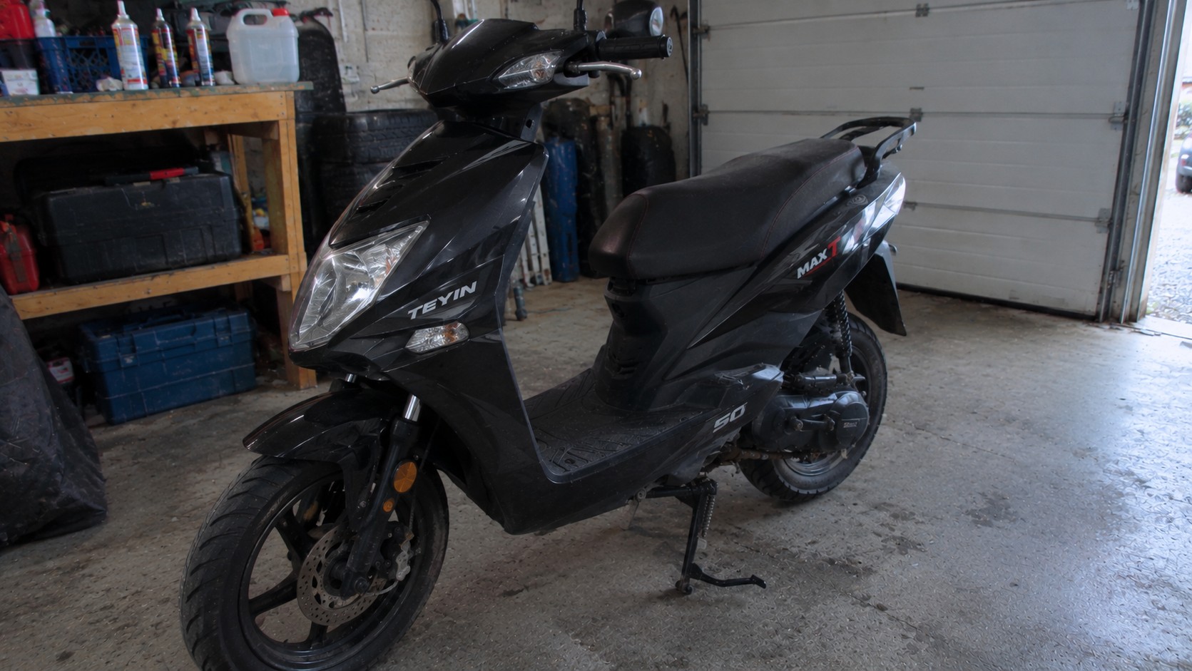 teyin_max_t_50cc_scooter_noir_vue_trois_quarts_avant_carénage_sportif_phare_avant_guidon_rétroviseurs_selle_noire_porte_bagages_roue_avant_frein_à_disque_fourche_avant_suspension_arrière_moteur_transmission_carter_50cc_béquille_centrale_sol_béton_garage_francais_établi_outils_bacs_rangement_pneus_porte_de_garage_sectionnelle_lumière_naturelle_ambiance_atelier_2026_10_07