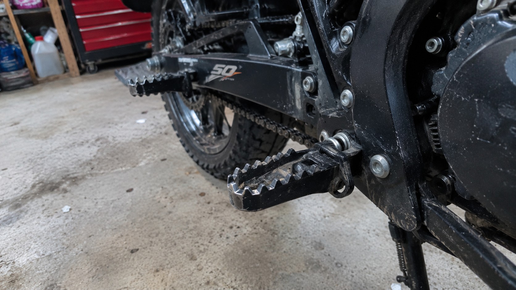 cale-pied_moto_50cc_noir_en_metal_crante_avec_dents_antiderapantes_axe_de_pivot_ressort_de_rappel_et_boulonnerie_de_fixation_sur_cadre_noir_vue_rapprochee_cote_moteur_chaine_transmission_bras_oscillant_pneu_arriere_sol_beton_tache_garage_2026-10-11