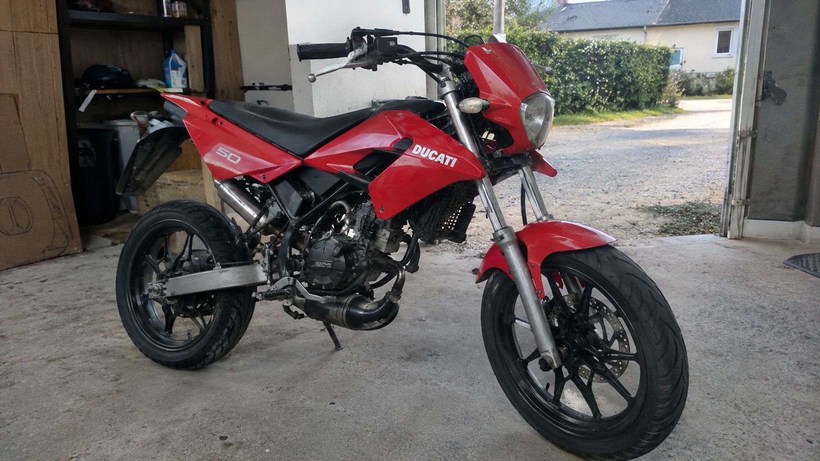 Ducati_50cc_rouge_vue_trois_quarts_avant_gauche_carénages_rouges_logo_Ducati_blanc_numero_50_selle_noire_moteur_thermique_noir_pot_echappement_argente_fourche_avant_argentee_jantes_noires_multibranches_pneus_route_frein_a_disque_bequille_laterale_garage_maison_francaise_sol_beton_etageres_outillage_lumiere_naturelle_ensoleillee_photo_amateur_realiste_Panasonic_GH4_2026-10-16